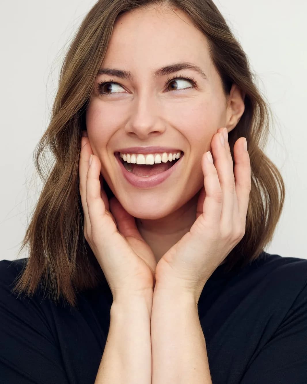 A delighted woman in a black top smiling with her hands on her cheeks